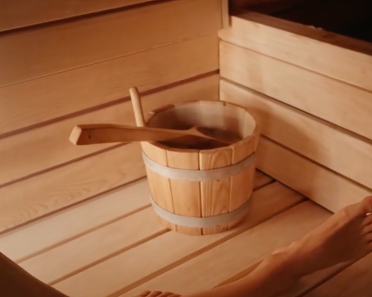 Person relaxing in a wooden sauna with a wooden bucket and ladle.